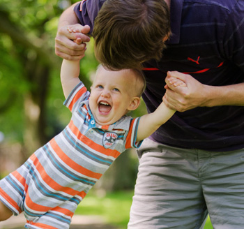 Father makes his toddler son laugh by swinging him while photographer takes a photo